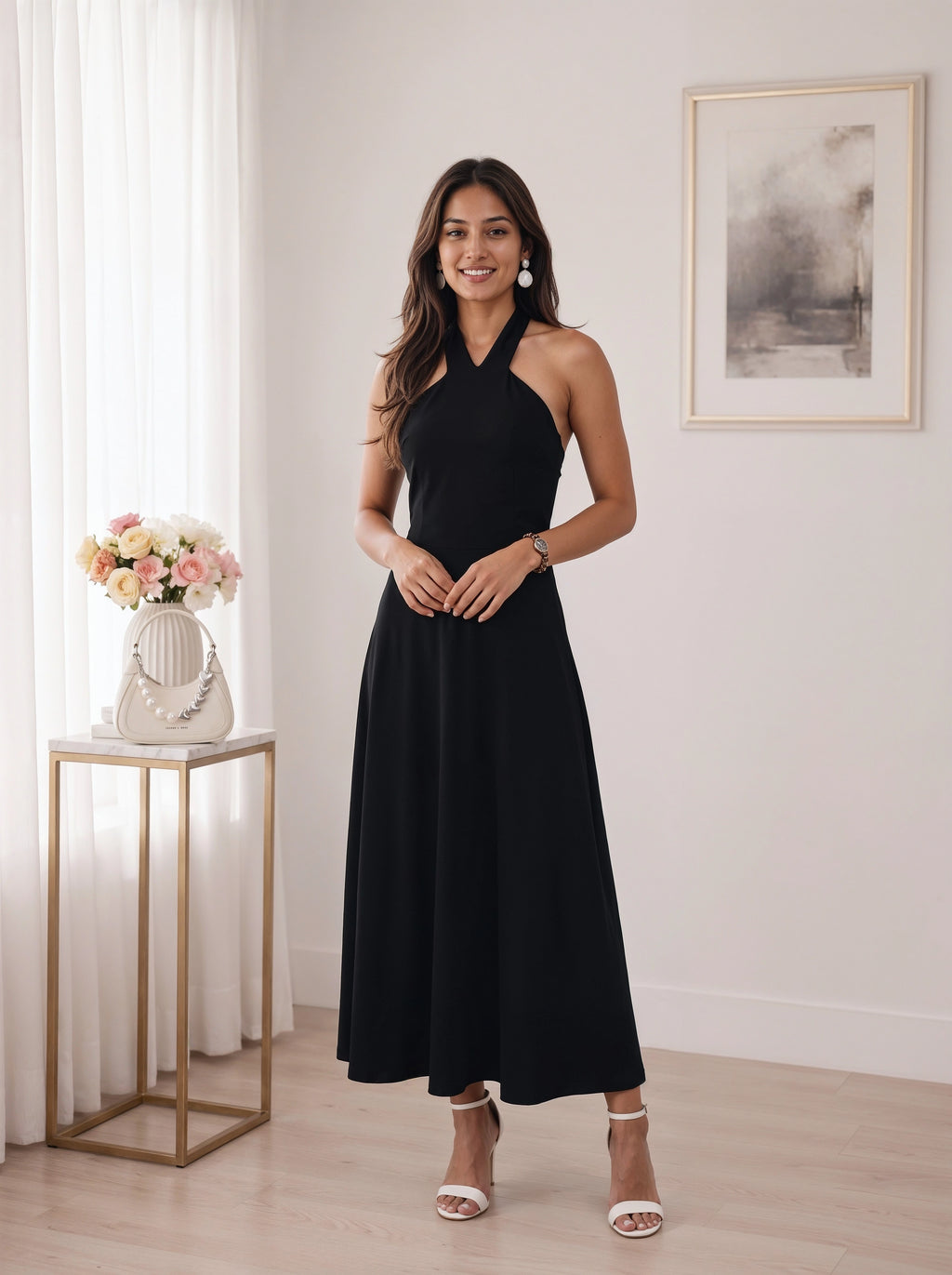 A woman modeling a sleeveless black halter-neck maxi dress in a bright, modern room with white curtains, a gold side table with flowers, and a framed print on the wall.