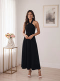 A woman modeling a sleeveless black halter-neck maxi dress in a bright, modern room with white curtains, a gold side table with flowers, and a framed print on the wall.