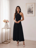 A woman modeling a sleeveless black halter-neck maxi dress in a bright, modern room with white curtains, a gold side table with flowers, and a framed print on the wall.