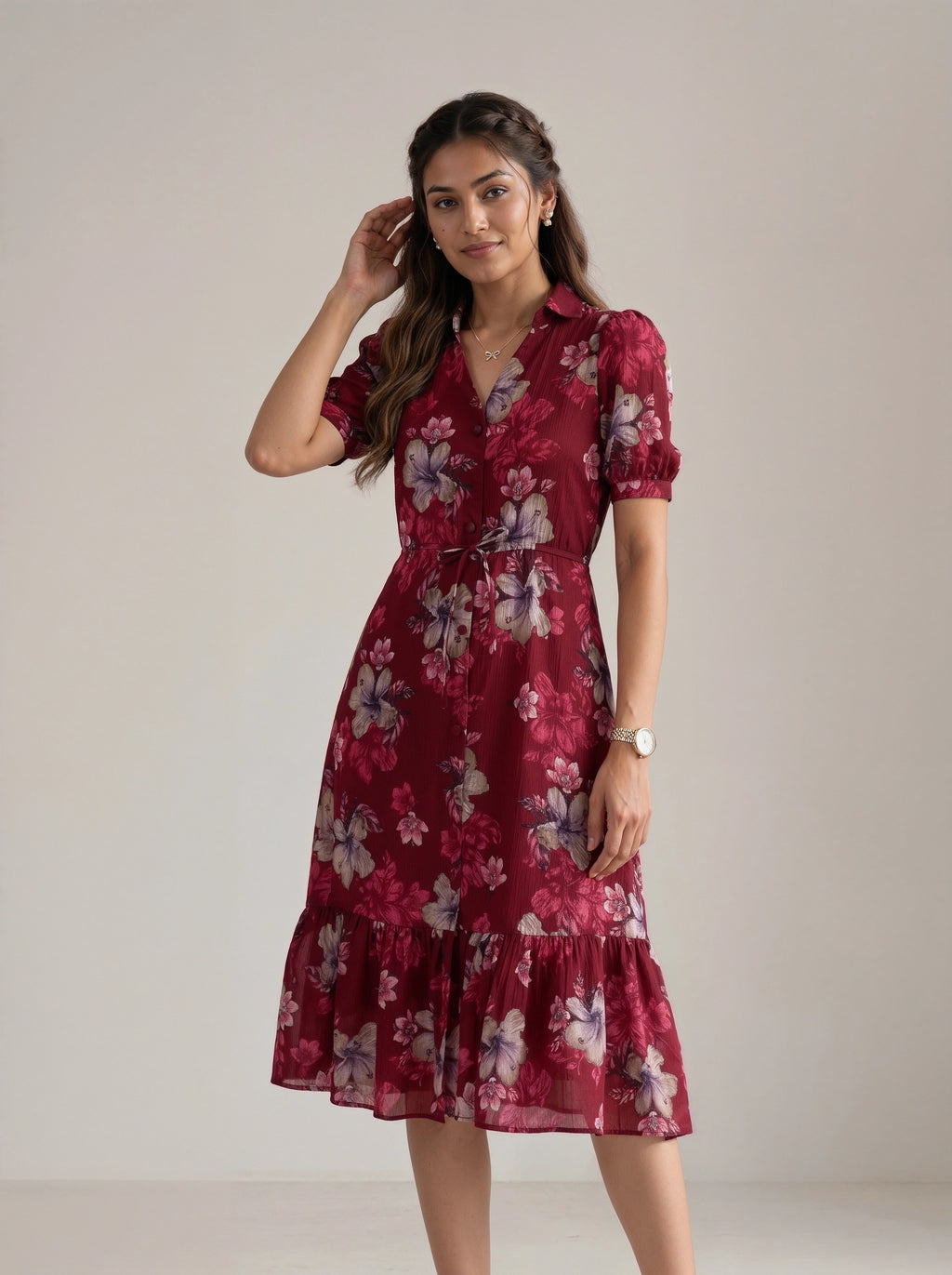 A woman modeling a short-sleeved midi dress with a floral print in deep red, burgundy, and pink, featuring a collared neckline, a tie waist, and a ruffled hem, set against a plain light beige studio background.