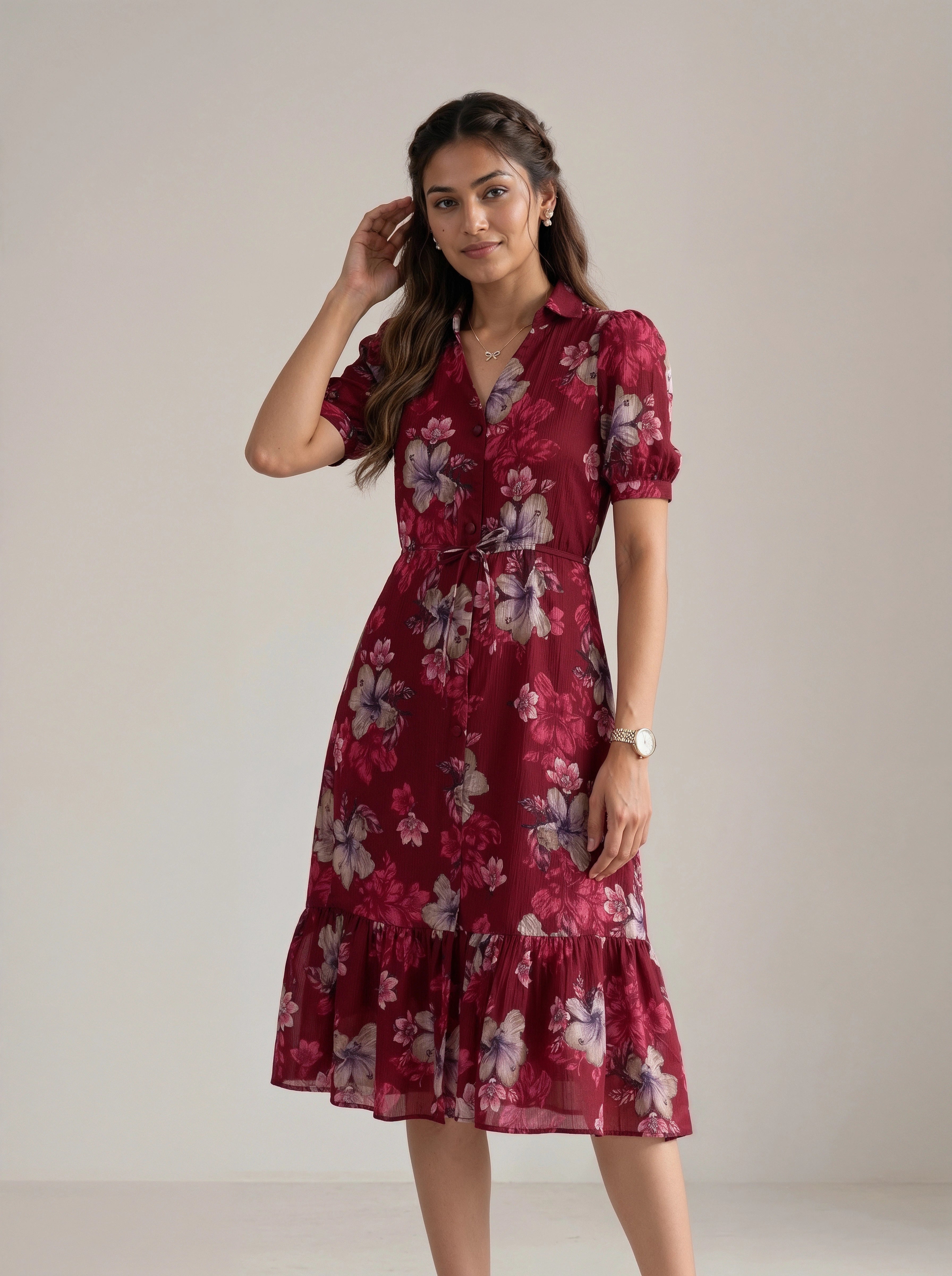 A woman modeling a short-sleeved midi dress with a floral print in deep red, burgundy, and pink, featuring a collared neckline, a tie waist, and a ruffled hem, set against a plain light beige studio background.