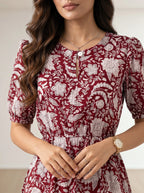 A woman wearing a short-sleeve red dress with a white floral toile pattern, featuring puff sleeves, a round neckline with a decorative keyhole button closure, and a gathered waist.