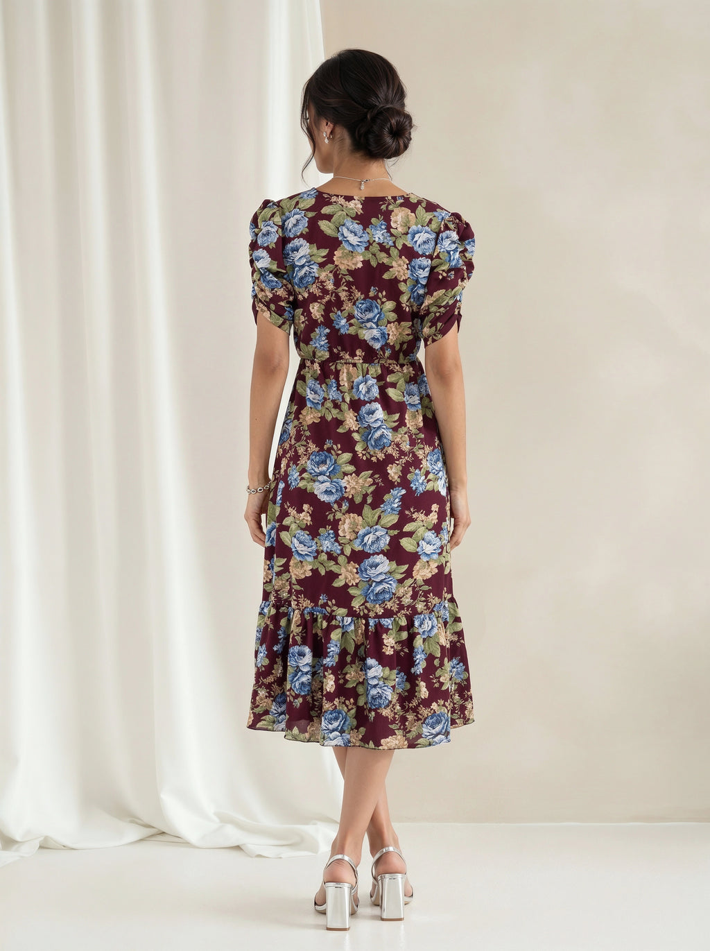 A woman viewed from behind wearing a short-sleeve midi dress with puff sleeves and a floral print on a dark burgundy background. The dress features blue, green, and beige flowers.