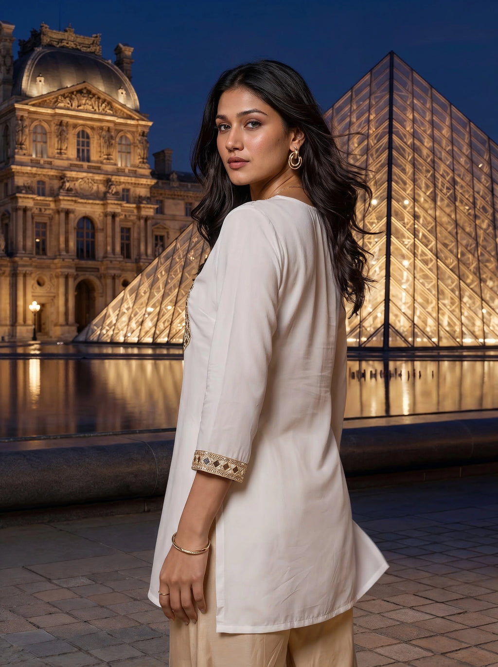 A woman wearing a cream-colored kurta with gold embroidered cuffs, standing in front of the illuminated Louvre Museum and glass pyramid at night.