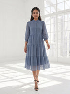 Woman wearing a blue dress standing in a bright room with large windows.