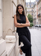 A woman posing on a city sidewalk next to a beige stone wall, wearing a black sleeveless top and black wide-leg trousers, with a white handbag resting on the wall beside her.