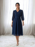 Woman wearing a navy blue dress standing in front of white curtains.