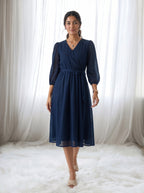 Woman wearing a navy blue dress standing in front of white curtains.
