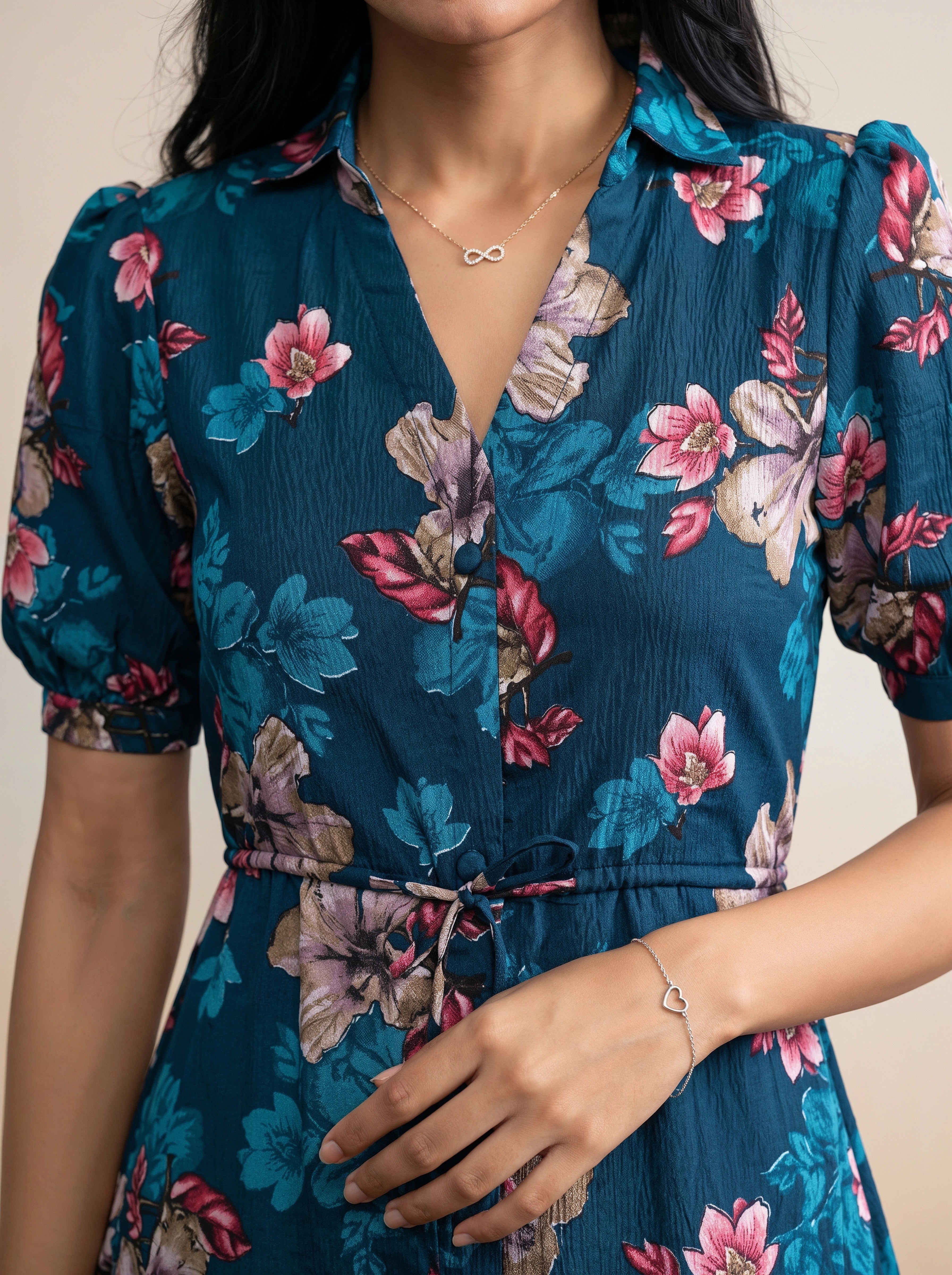 A close-up of a woman wearing a short-sleeve teal blue floral dress with pink, purple, and green flowers, featuring a tie waist and a V-neckline.