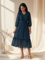 Woman wearing a blue dress standing in a softly lit room with curtains.