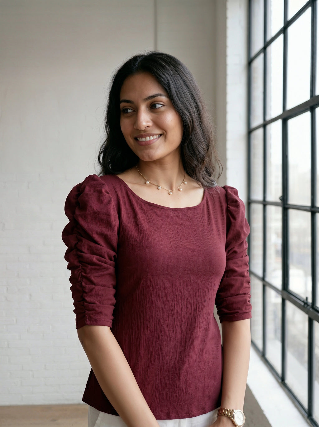 Woman wearing a burgundy top with puffed sleeves standing by a window.
