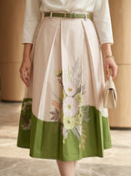 Woman wearing a white blouse and floral skirt with a green belt, holding a beige handbag.