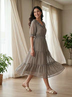 Woman wearing a gray dress standing in a room with curtains and plants.