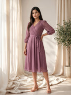 Woman wearing a purple dress standing in a softly lit room with curtains and a plant.