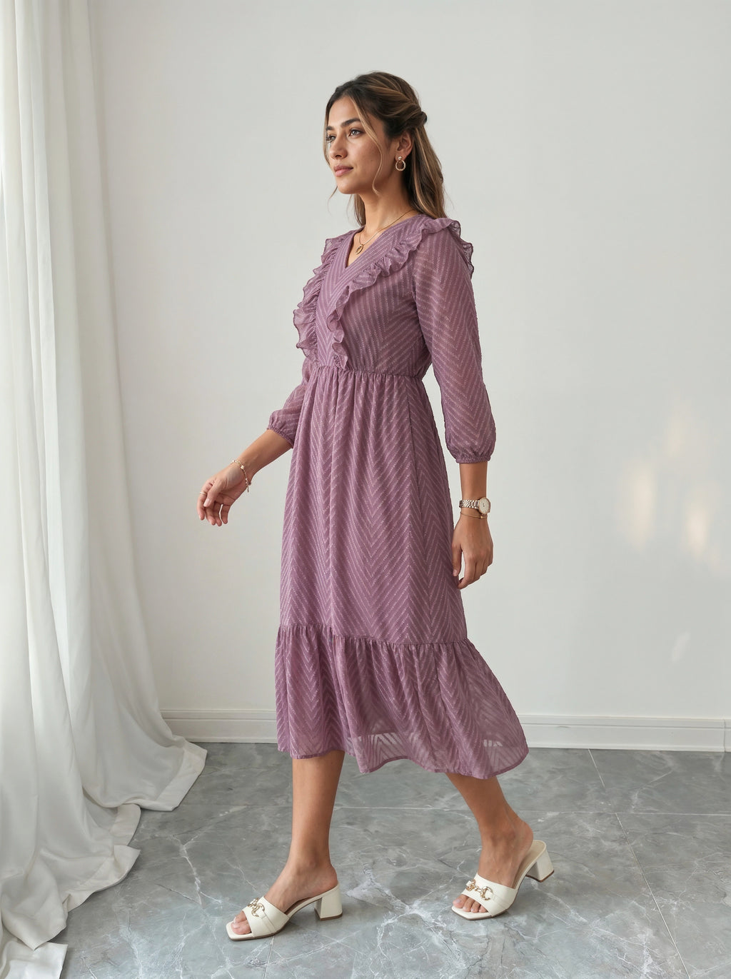 Woman wearing a purple dress standing in a room with white curtains and gray floor.