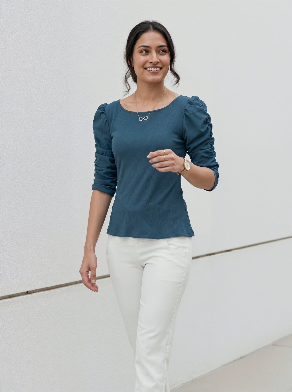 Woman wearing a teal top with puffed sleeves and white trousers standing against a neutral wall.