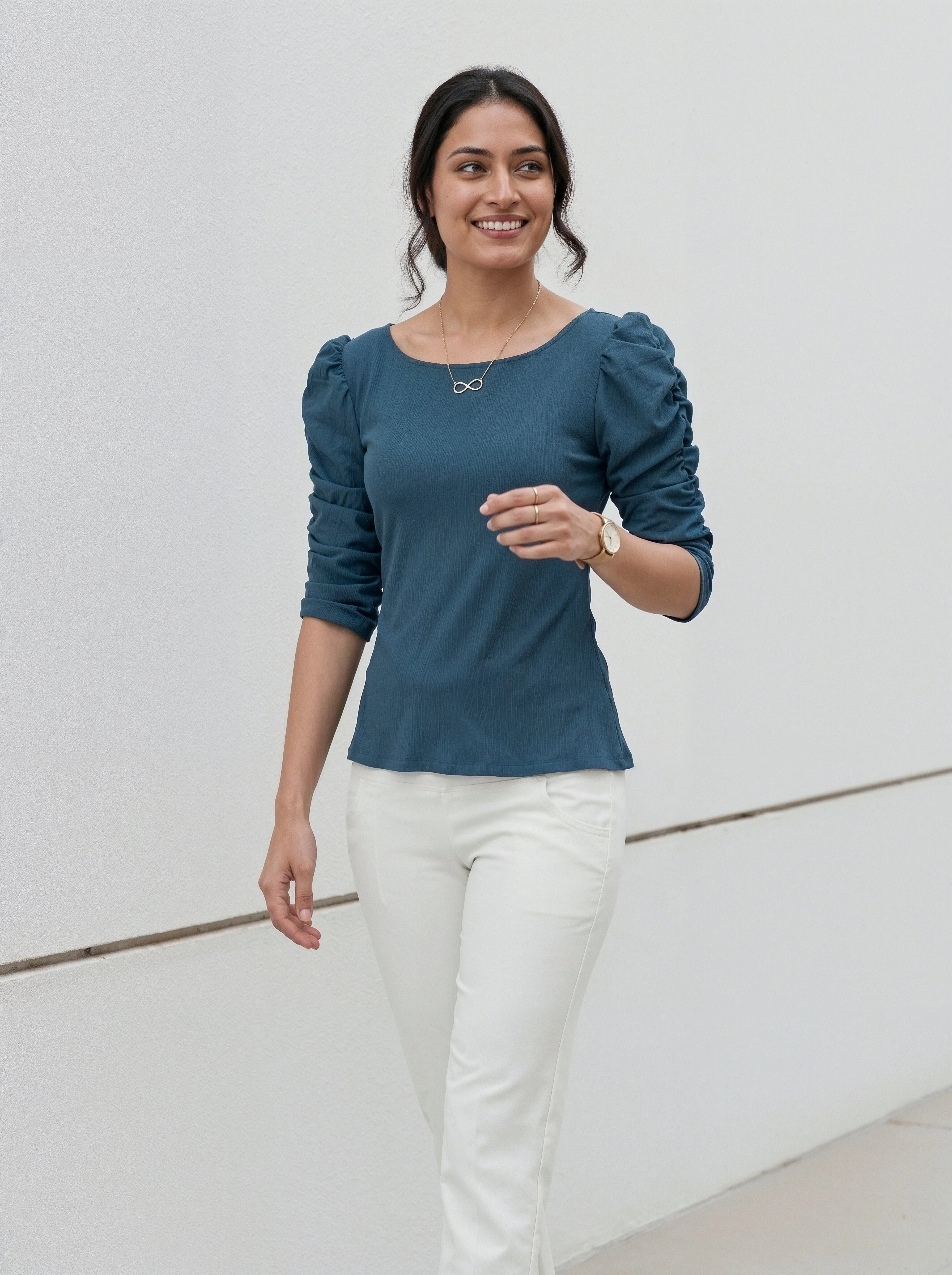 Woman wearing a teal top with puffed sleeves and white trousers standing against a neutral wall.