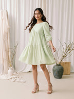 Woman wearing a light green dress standing in a softly lit room with white curtains and decorative vases.