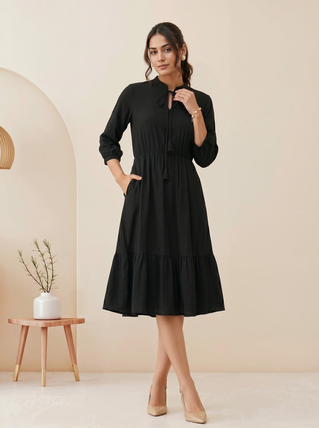 Woman wearing a black dress standing in a minimalistic room with a beige wall and wooden side table.