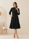 Woman wearing a black dress standing in a minimalistic room with a beige wall and wooden side table.