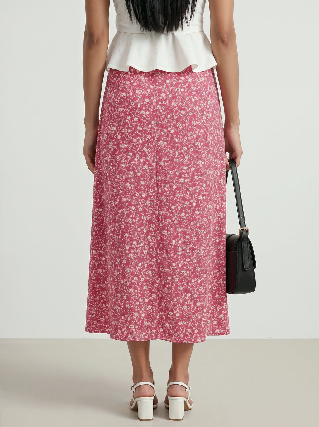Woman wearing a white top and pink floral skirt with a black handbag on a plain background