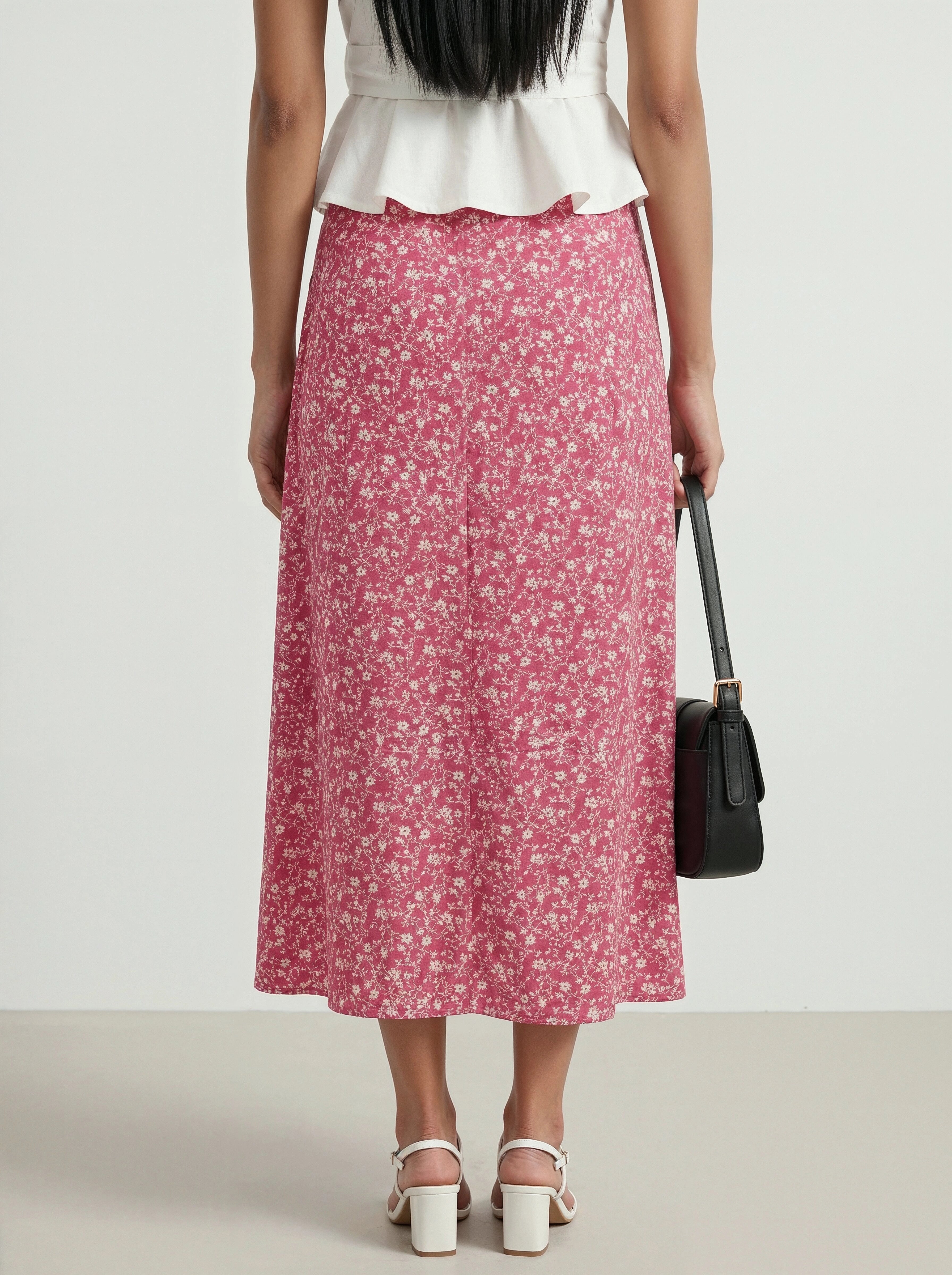 Woman wearing a white top and pink floral skirt with a black handbag on a plain background