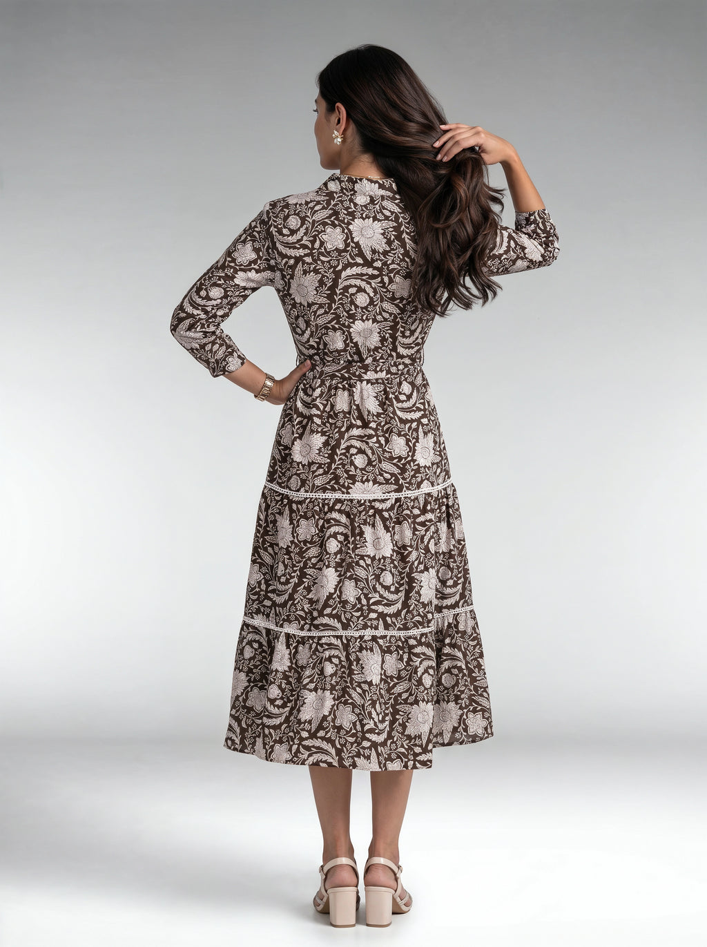 A woman is shown from the back wearing a long-sleeved midi dress with a brown and beige floral paisley pattern, paired with beige block-heel sandals, against a light gray studio background.