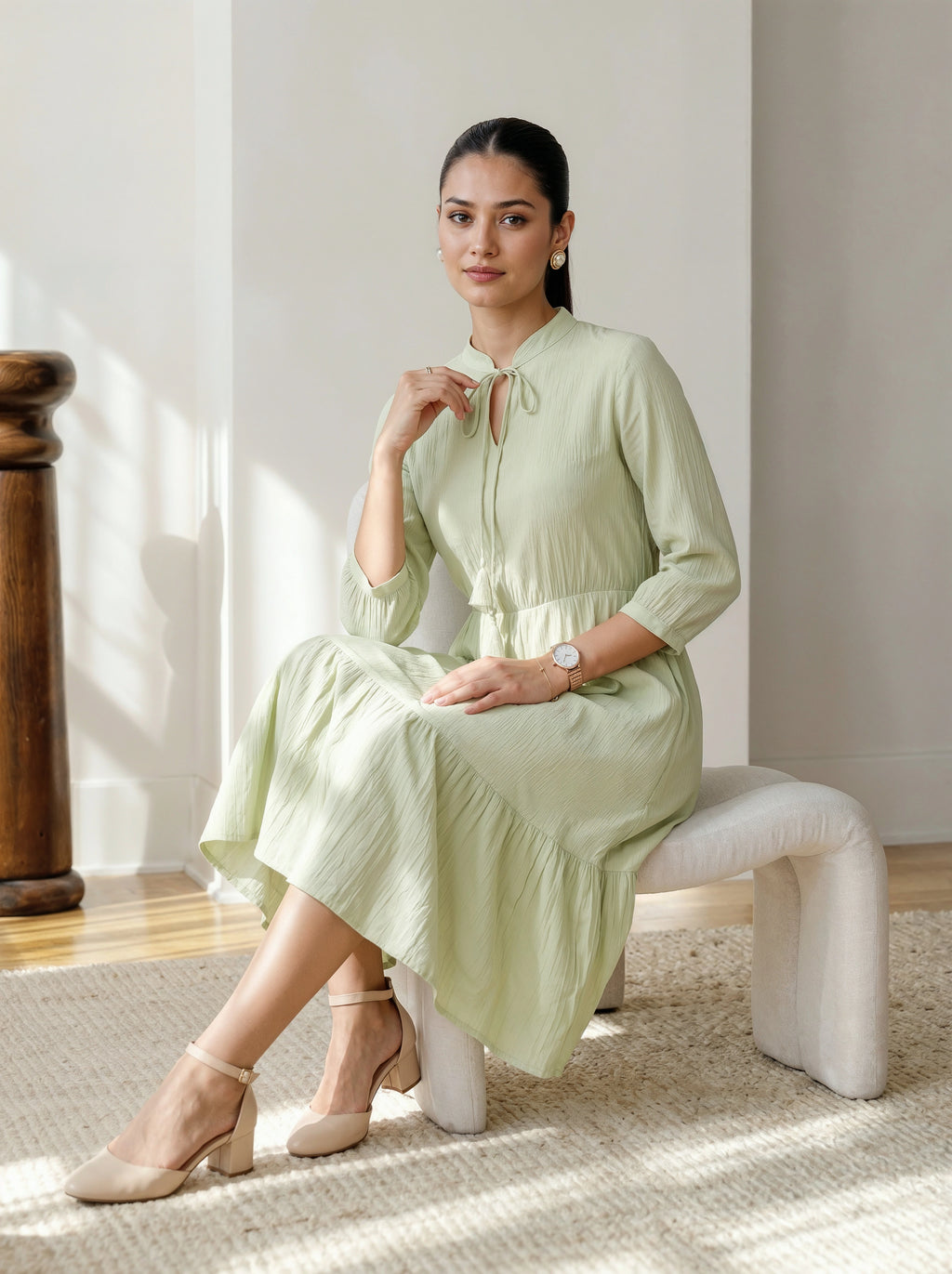 Woman in a light green dress sitting on a white stool in a bright room.