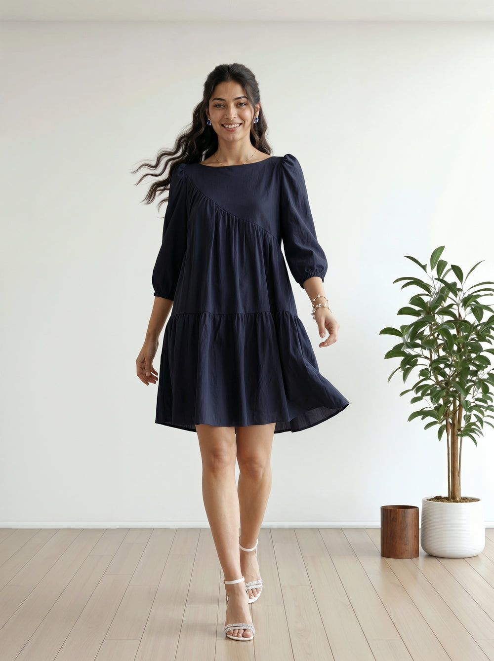 Woman wearing a navy blue dress standing in a room with a white wall and a plant.