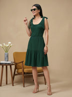 Woman in a green dress standing in a room with a chair and table in the background.