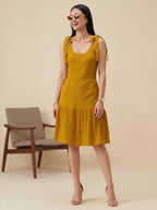 Woman wearing a mustard yellow dress standing in front of a beige background with a chair.