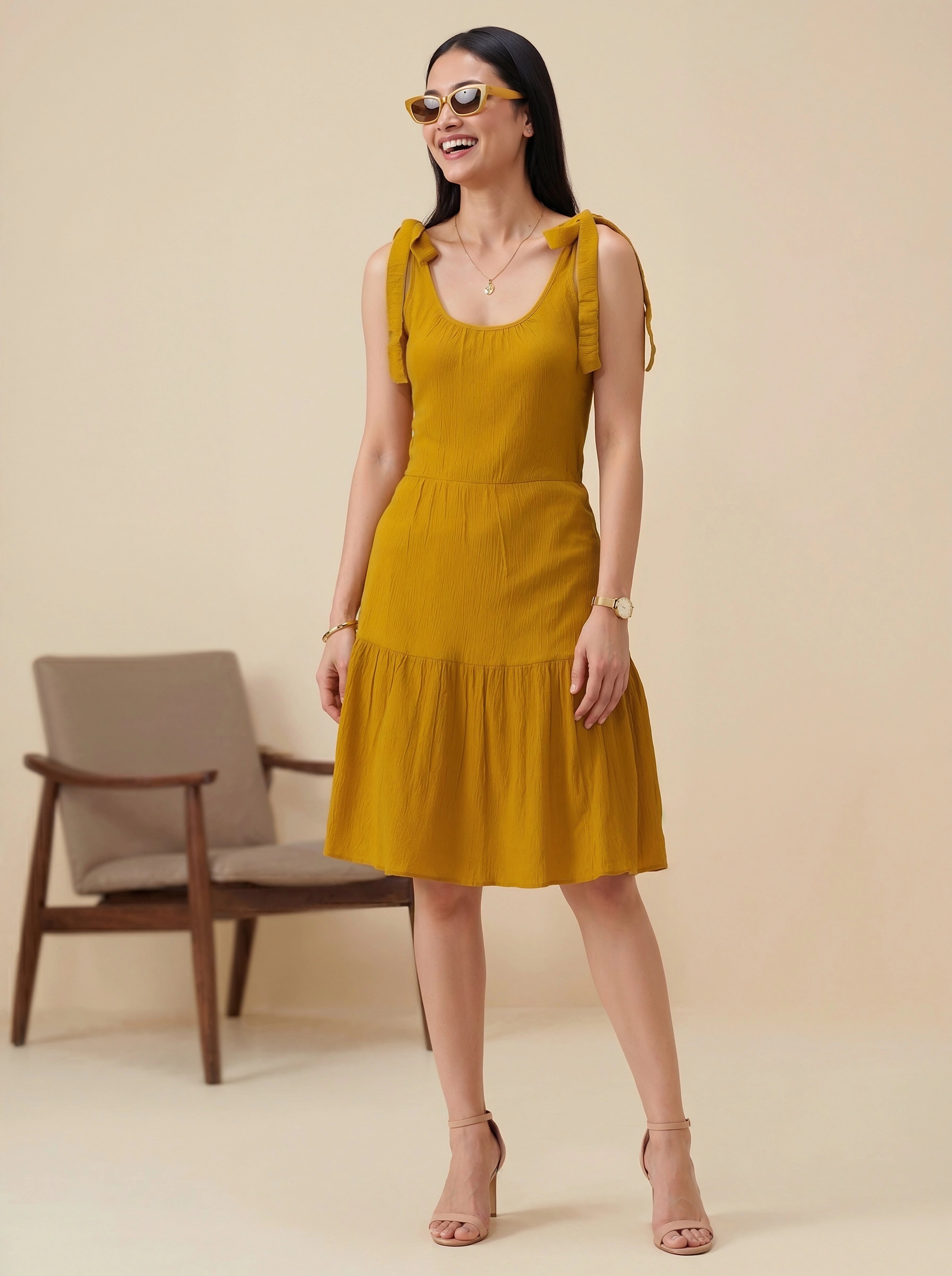 Woman wearing a mustard yellow dress standing in front of a beige background with a chair.