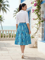 Woman in a white blouse and blue patterned skirt standing in front of a stone building with palm trees and ocean view.