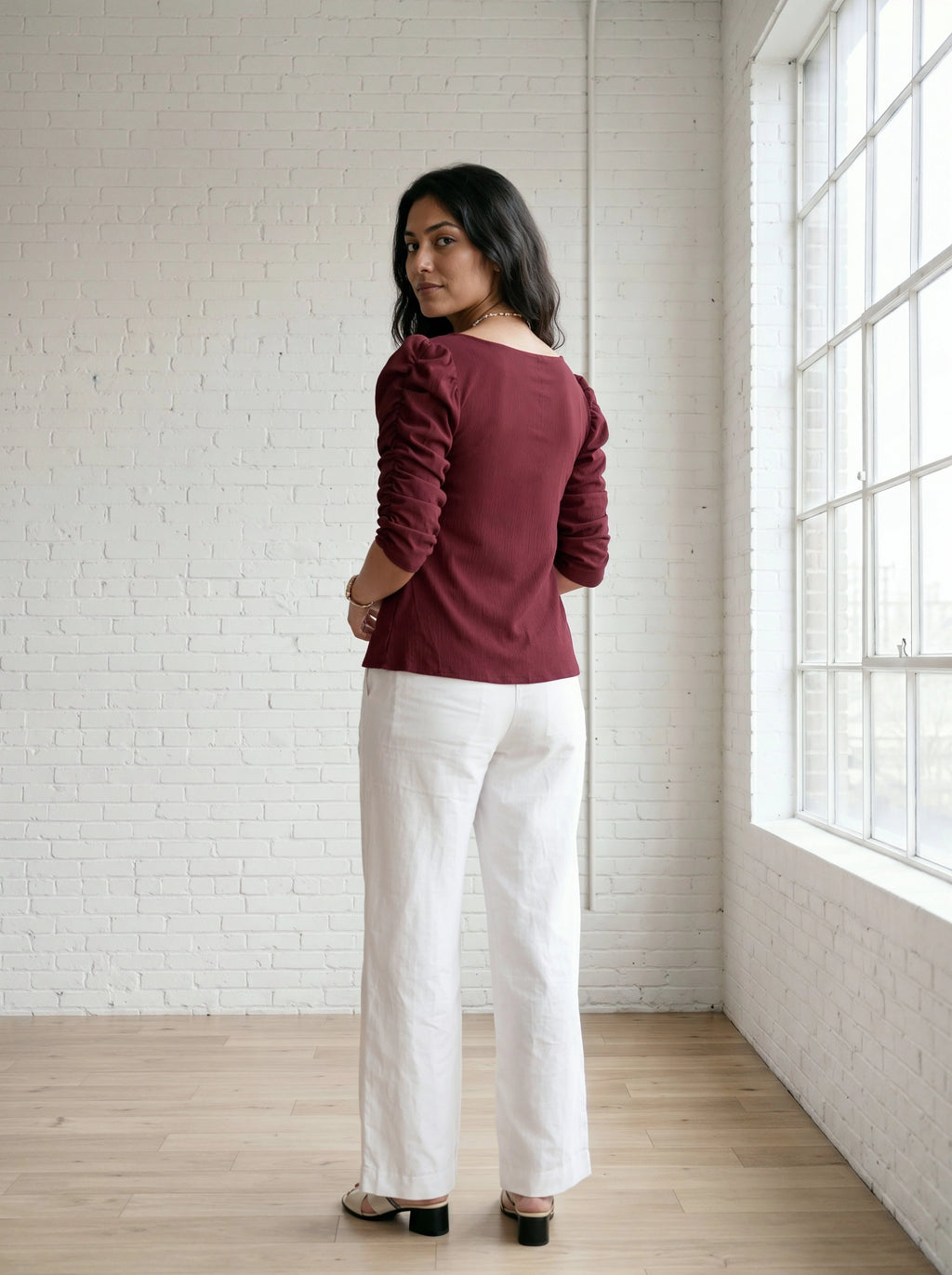 Woman wearing a burgundy top and white pants standing in a room with a white brick wall and large window.
