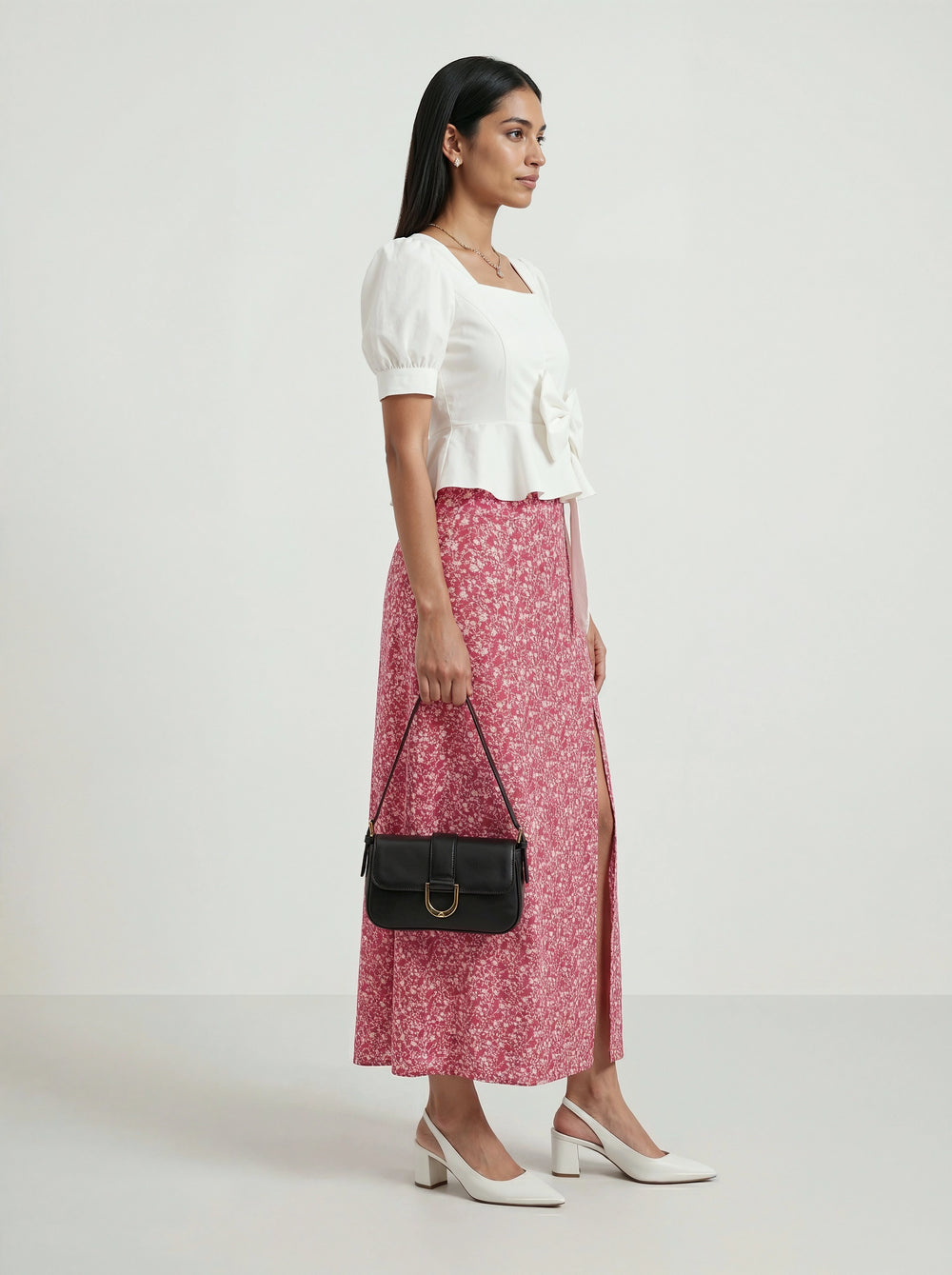 Woman wearing a white top and pink floral skirt, holding a black handbag on a white background