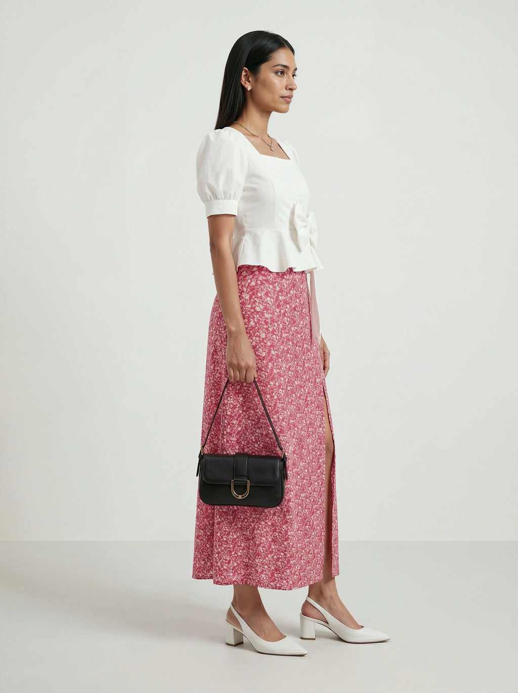 Woman wearing a white top and pink floral skirt, holding a black handbag on a white background
