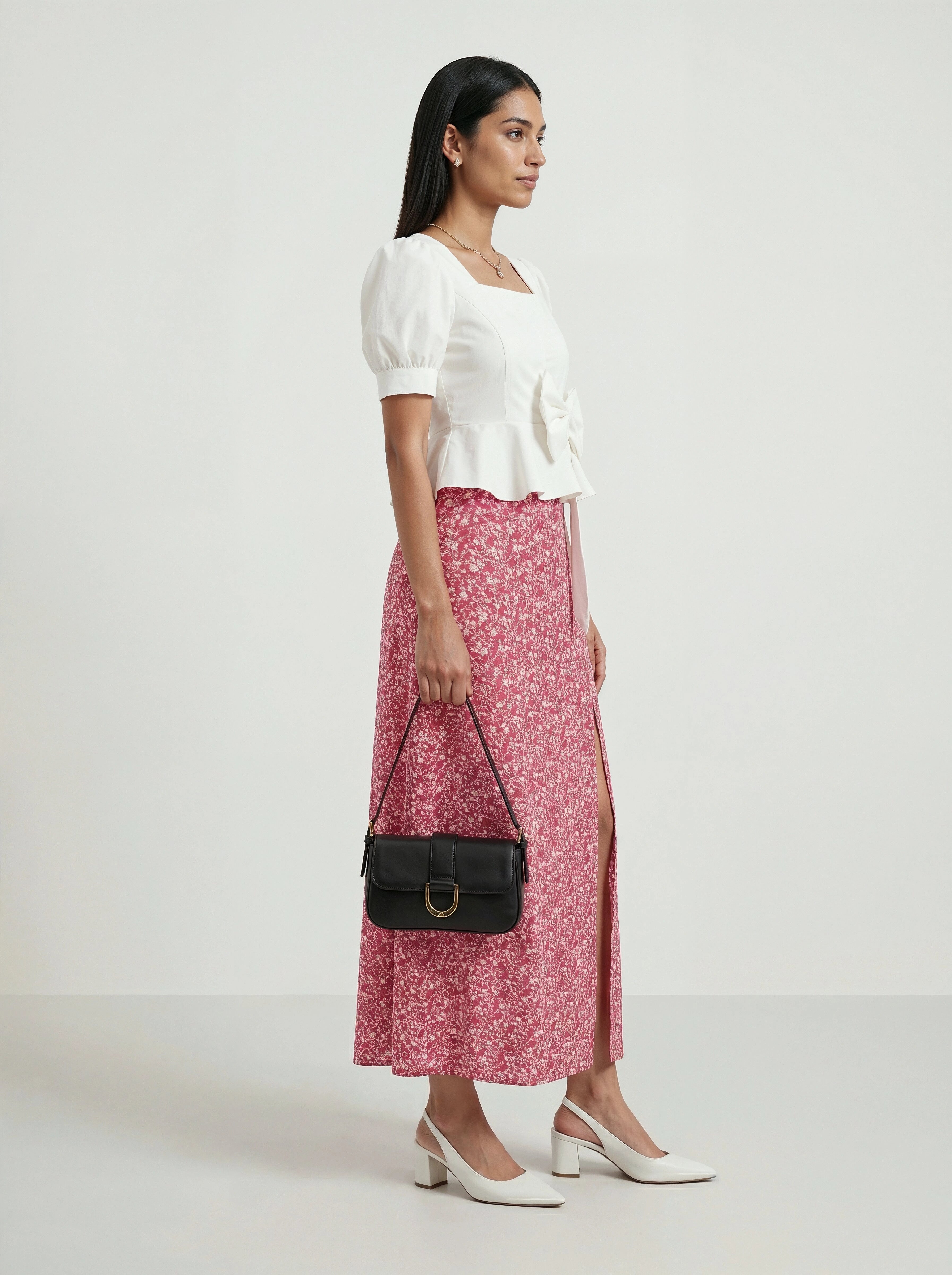 Woman wearing a white top and pink floral skirt, holding a black handbag on a white background