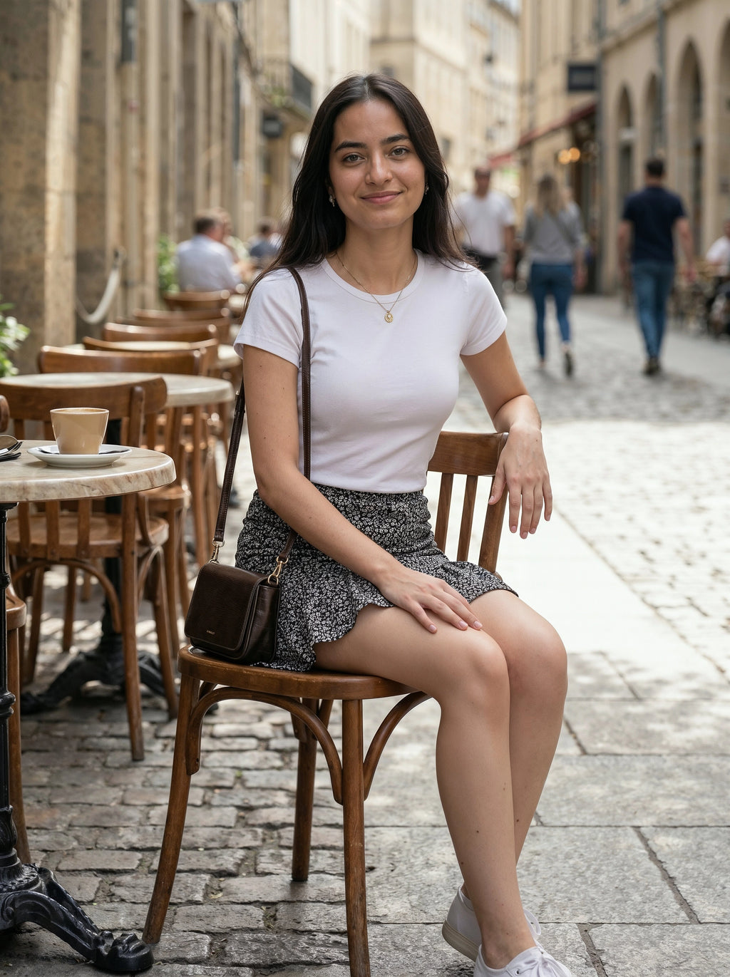 Woman sitting on a wooden chair in an outdoor cafe setting with people and buildings in the background.