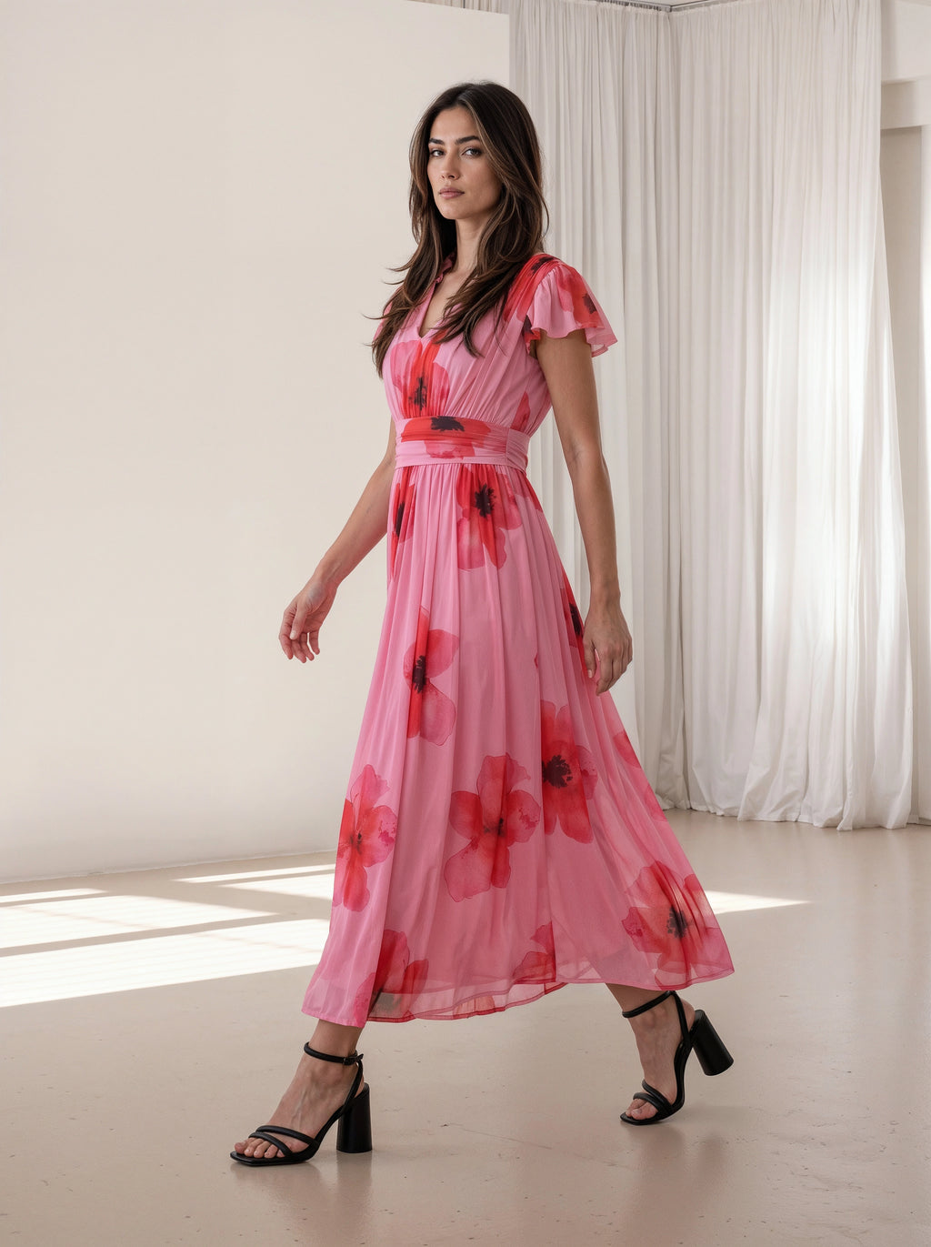 A woman modeling a pink midi dress with a red and black floral print, featuring short flutter sleeves and a matching waist belt, in a bright studio setting with white curtains.