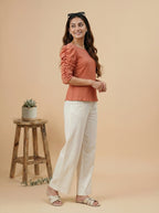 Woman wearing a rust-colored top and white pants standing next to a wooden stool with a plant on a beige background
