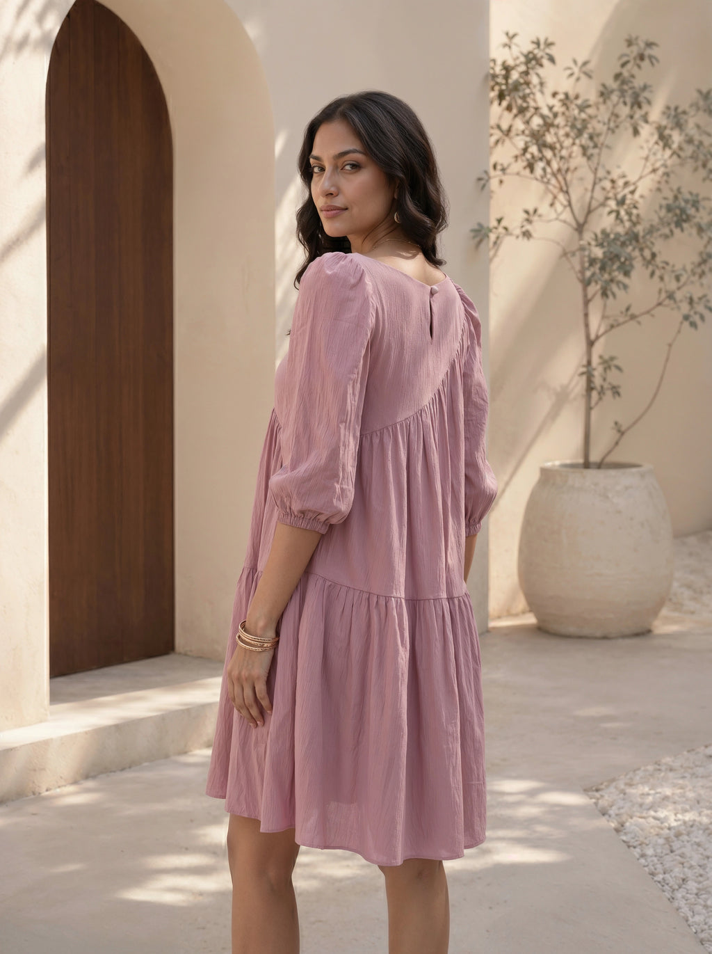 Woman in a pink dress standing in a sunlit outdoor setting with a wooden door and plant in the background.
