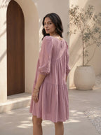 Woman in a pink dress standing in a sunlit outdoor setting with a wooden door and plant in the background.