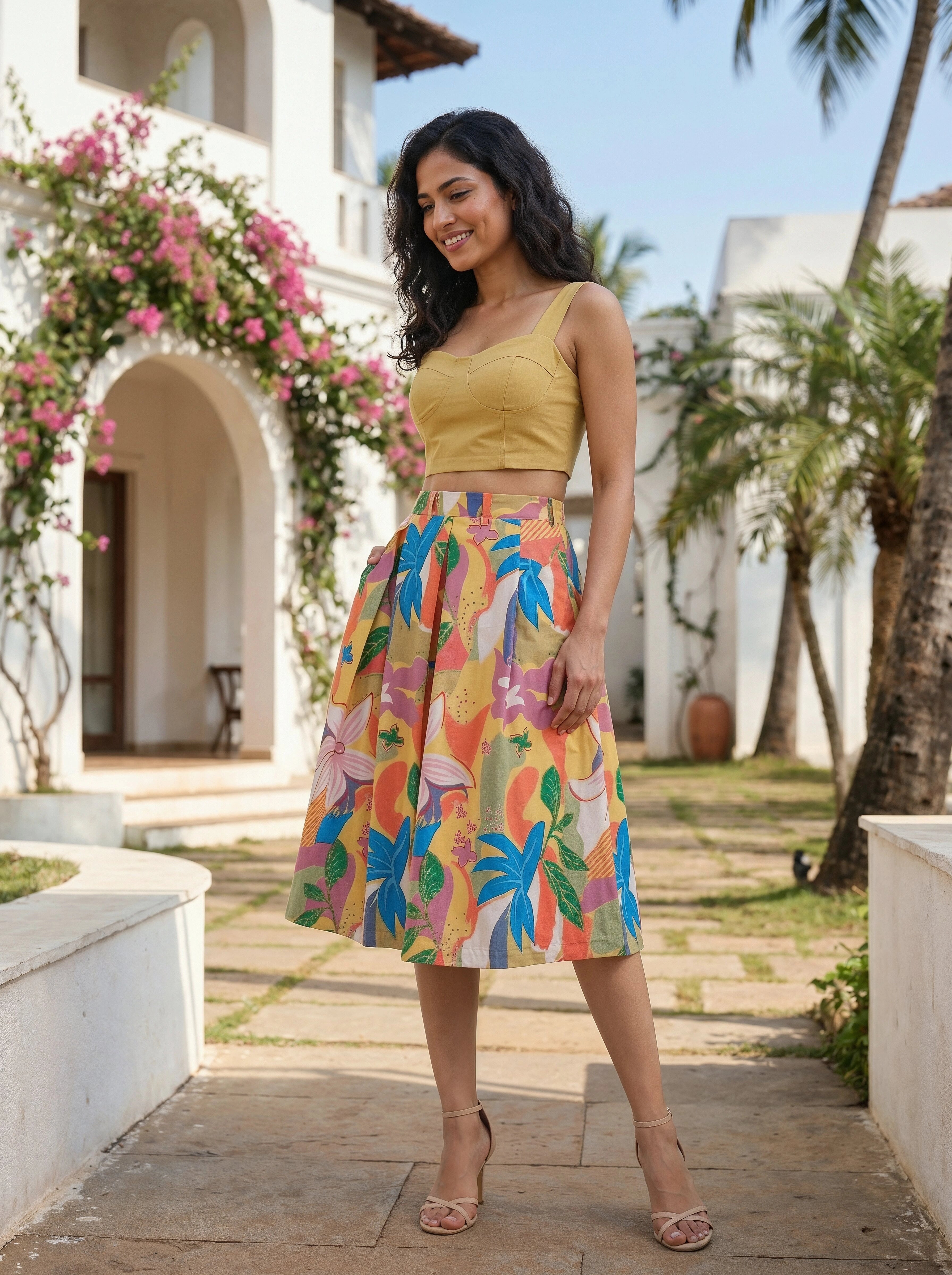 Woman in a yellow top and colorful skirt standing outdoors with floral decorations and a white building in the background.