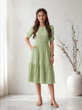 Woman wearing a light green dress standing in a bright room with a vase and plant in the background.