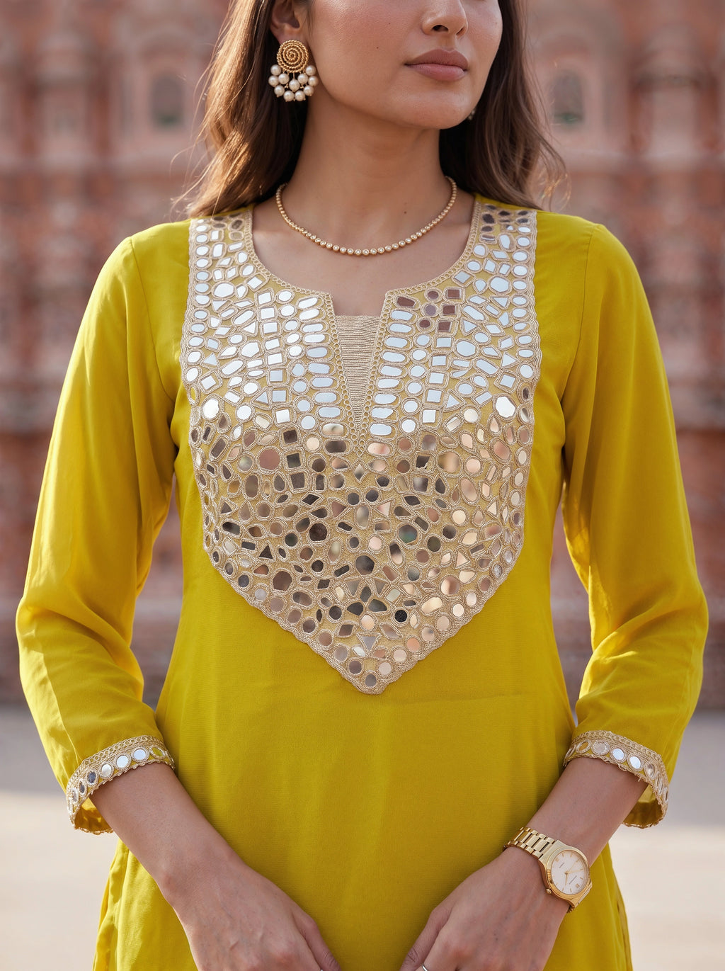A woman wearing a bright yellow kurta with extensive gold and silver mirror work embroidery on the chest and cuffs, posed outdoors against a soft-focus brick wall.