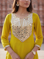 A woman wearing a bright yellow kurta with extensive gold and silver mirror work embroidery on the chest and cuffs, posed outdoors against a soft-focus brick wall.