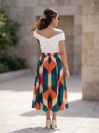 A woman photographed from behind wearing a white off-the-shoulder wrap top and a midi skirt with a bold abstract geometric pattern in teal, orange, cream, and navy, standing outdoors against a stone wall.