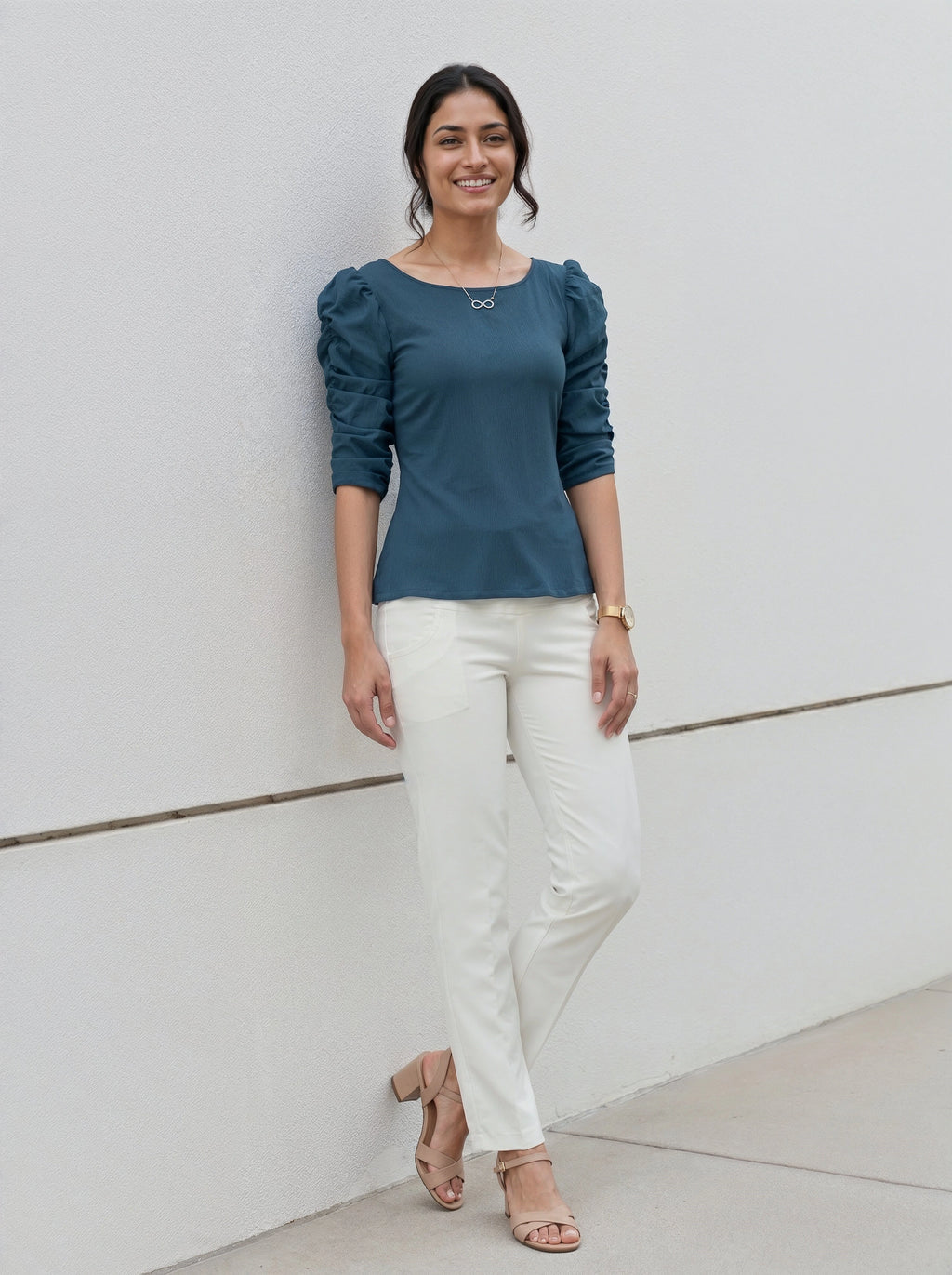 Woman wearing a teal puffed sleeve top and white pants standing against a light background