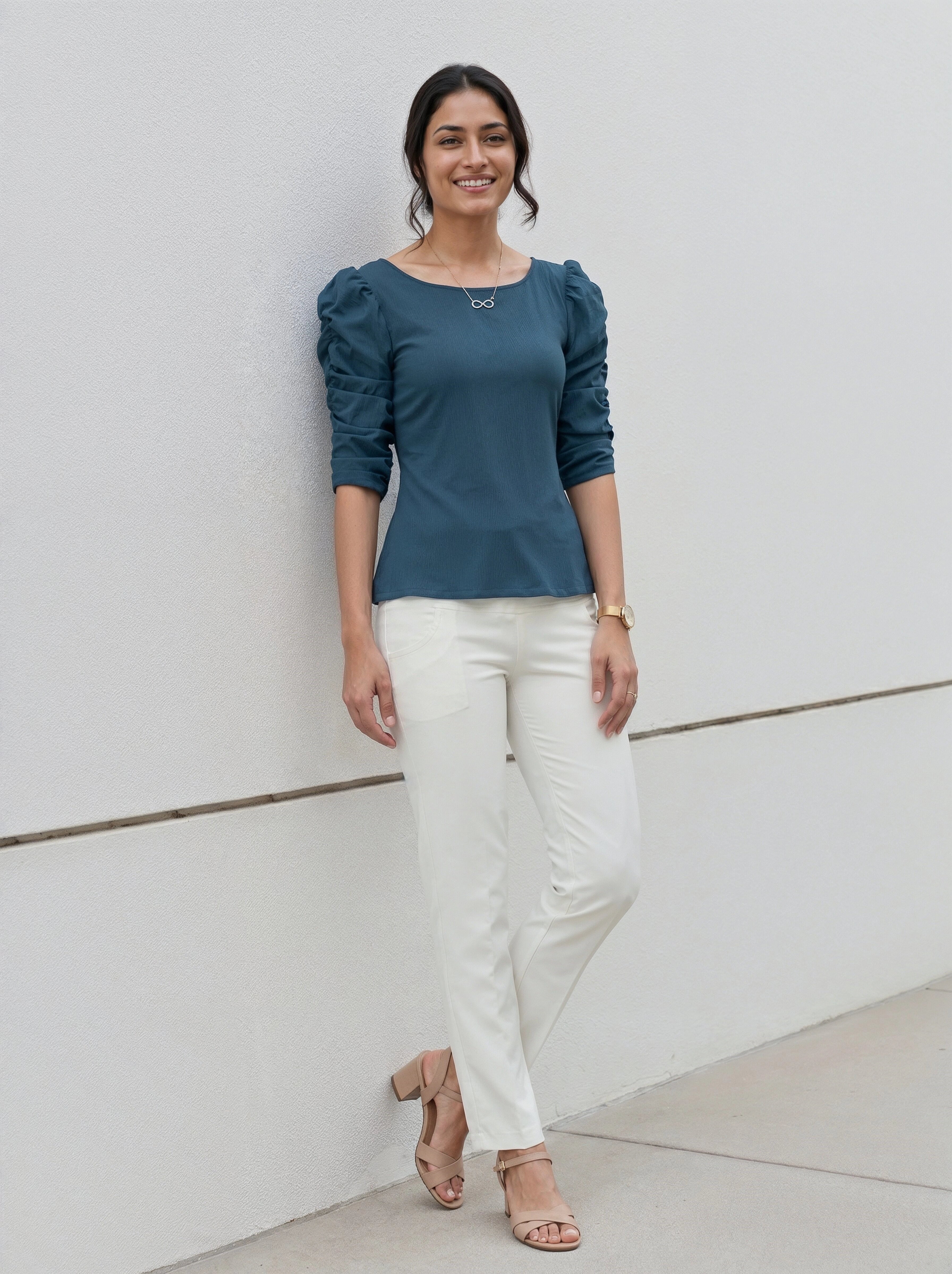Woman wearing a teal puffed sleeve top and white pants standing against a light background