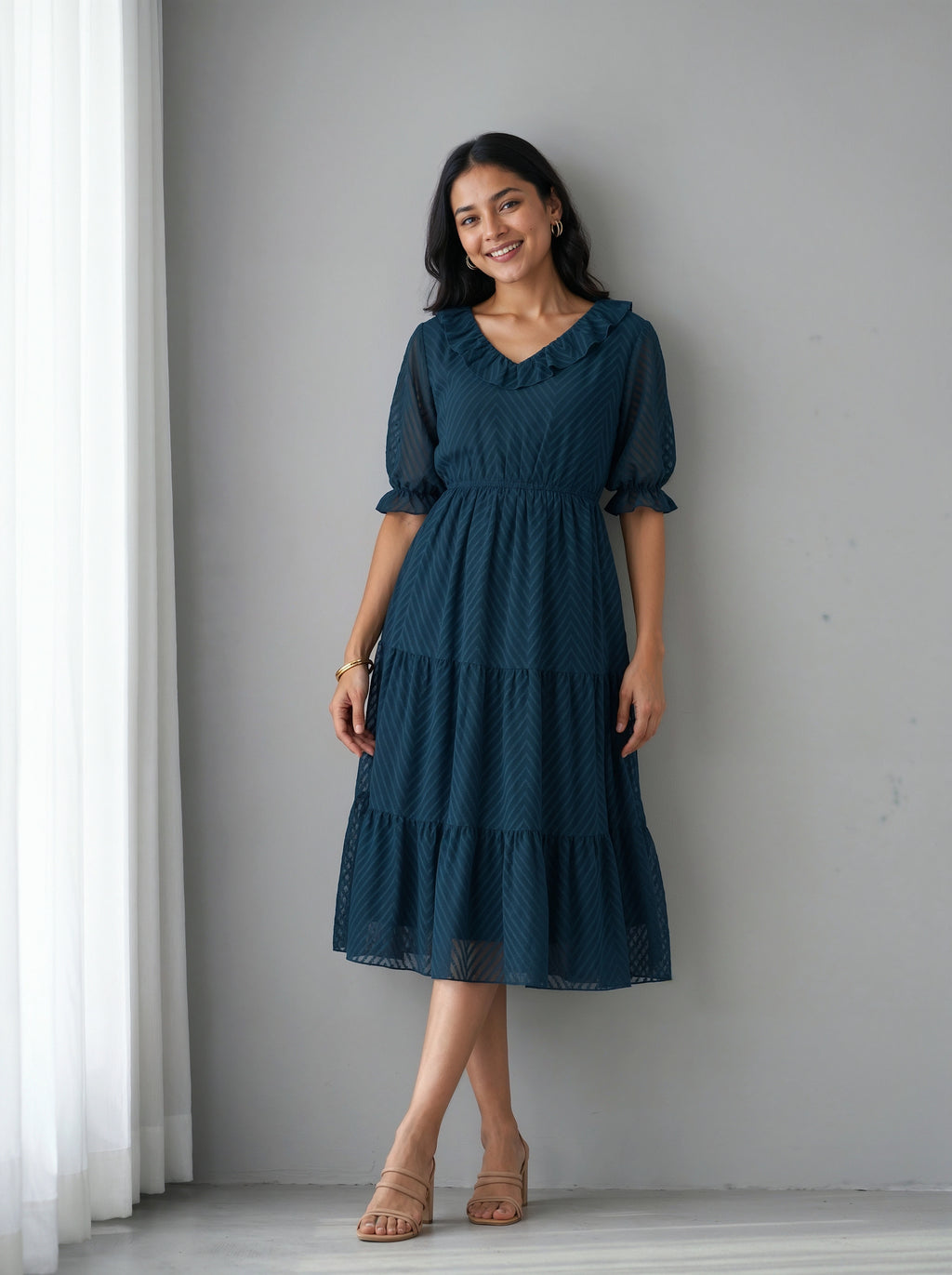 Woman wearing a blue dress standing against a gray wall with white curtains.